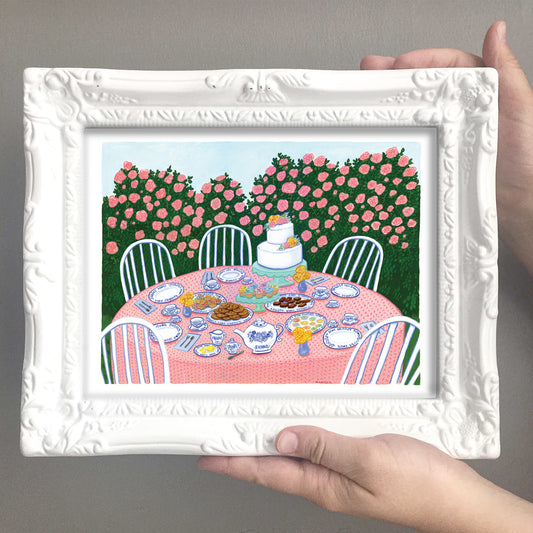 Framed artwork of a garden party scene with a table, chairs, and cake.
