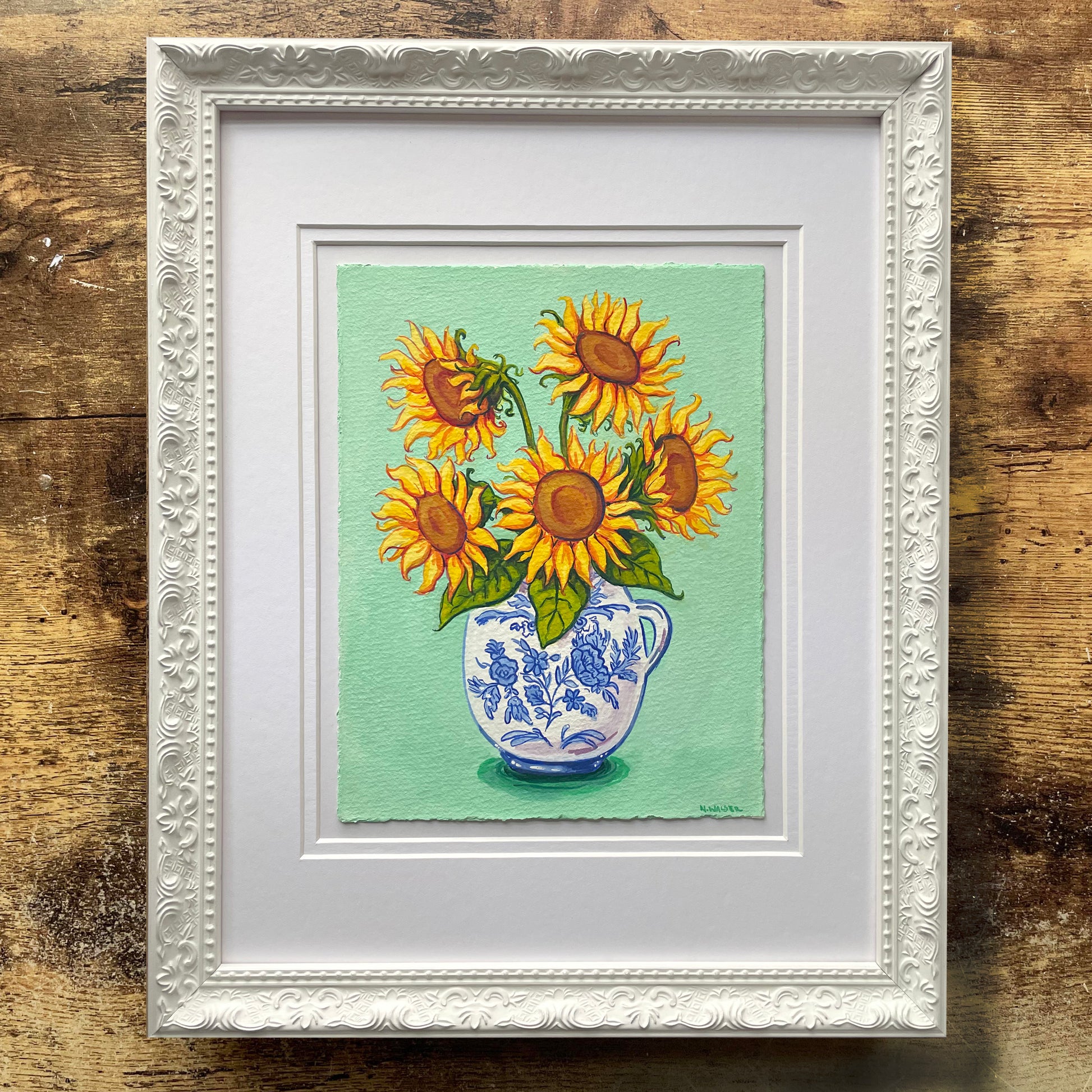 Framed artwork of sunflowers in a blue and white vase on a green background