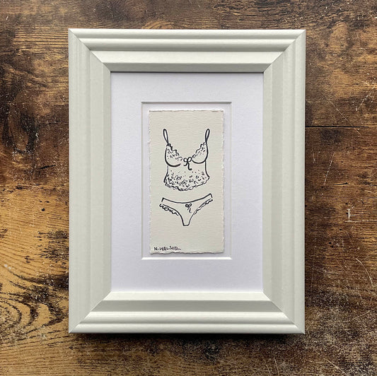 Framed black and white illustration of lingerie on a wooden surface