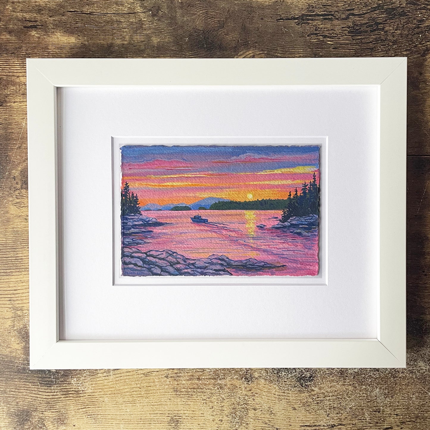 Framed artwork of a sunset over a lake with a boat, on a wooden surface.
