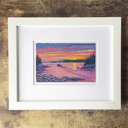 Framed artwork of a sunset over a lake with a boat, on a wooden surface.