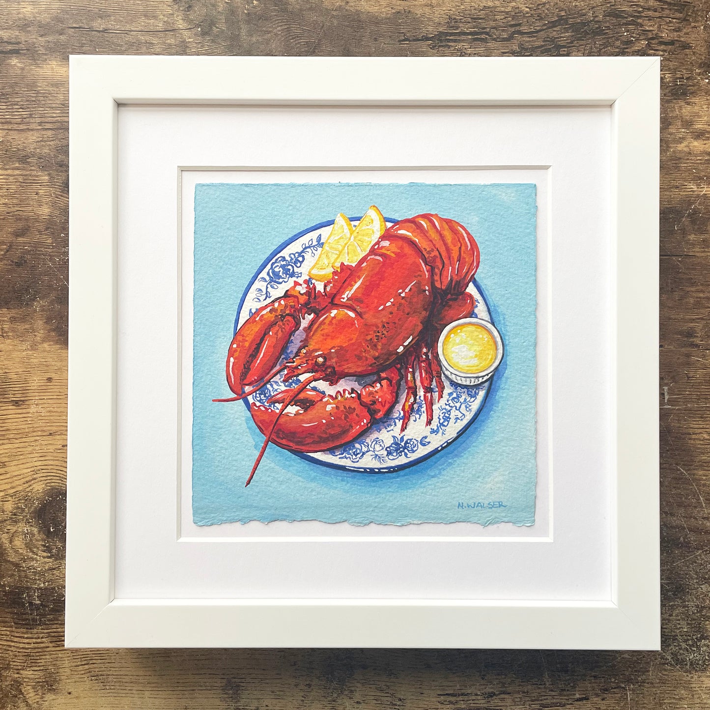 Framed artwork of a lobster on a plate with lemon wedges against a blue background.