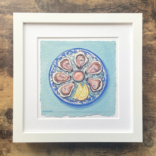 Framed artwork of oysters on a blue plate with lemon wedges on a textured wooden surface