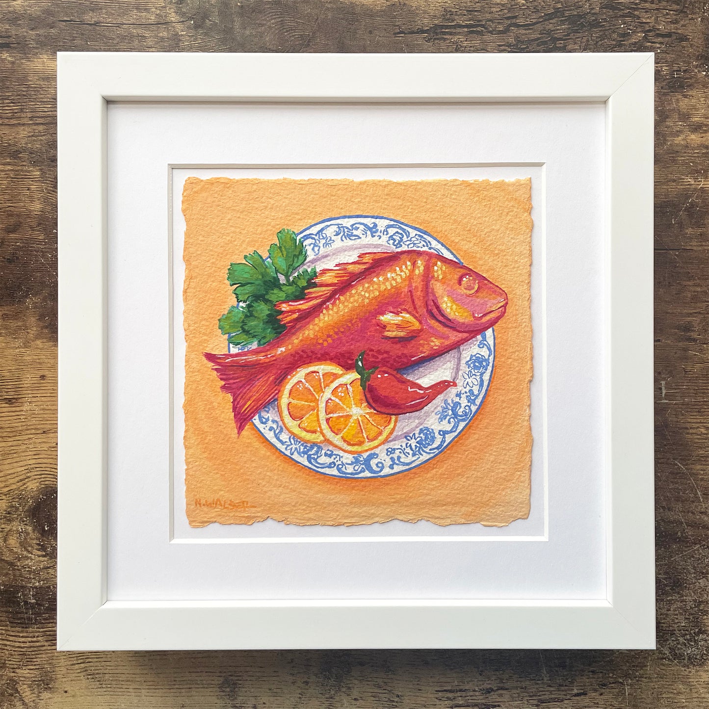 Framed artwork of a red fish on a plate with garnishes on an orange background